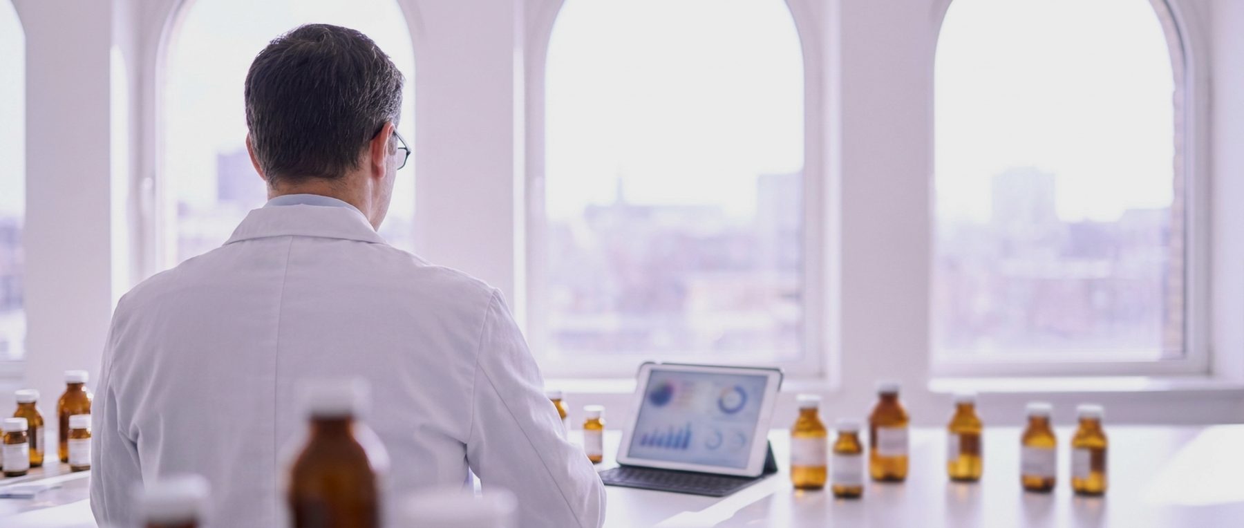 A clinical pharmacist seen from behind at a bright bench reviewing prescription vials and a tablet.