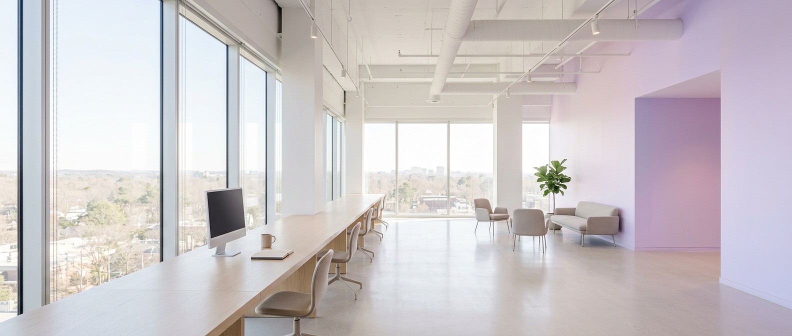 A bright, open plan office interior with tall windows and a long empty desk.