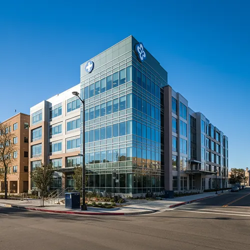 A modern, multi-story medical office building with large glass windows, a healthcare cross symbol on top, and a clear blue sky overhead. The street in front is empty and lined with young trees, reflecting a place dedicated to quality medical care.