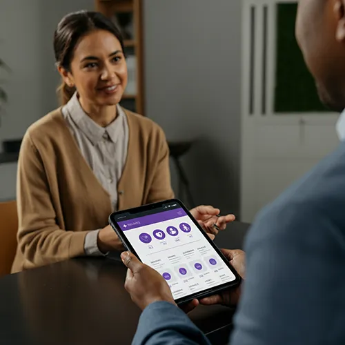 Two people sit across a table talking. One holds a tablet displaying health analytics on a purple-themed dashboard, while the other, wearing a brown cardigan, listens and smiles with care.