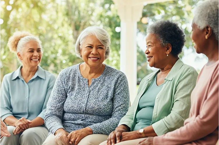 Four older women sit together outdoors, smiling and chatting in a sunny, green setting. Casually dressed, they appear to be enjoying each other’s company in a relaxed, friendly atmosphere that highlights the importance of social connections for health.