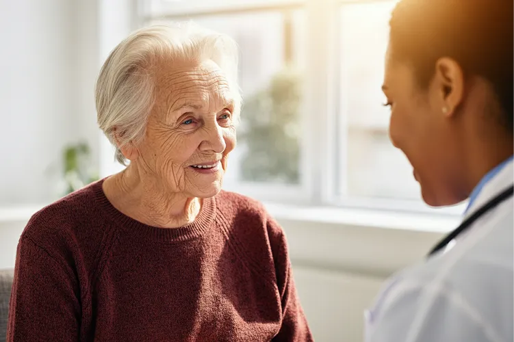 An elderly woman with gray hair and a maroon sweater smiles while talking to a healthcare professional in a white coat in a bright, sunlit room.