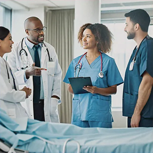 Four medical professionals, two men and two women, stand together in discussion near a hospital bed. They are smiling and wearing scrubs and lab coats, with one woman holding a clipboard.