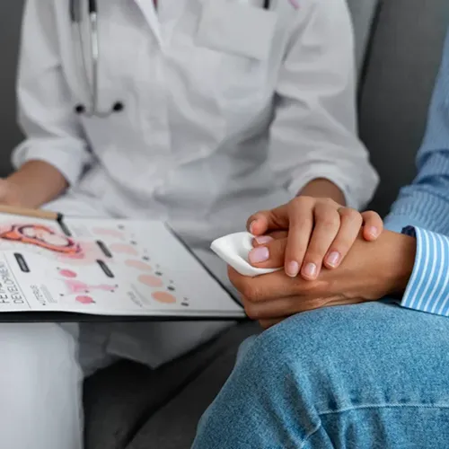 A doctor holding a medical chart sits with a patient. The patient, wearing blue, holds a tissue and is being comforted as they discuss test results. Only their hands and part of their bodies are visible.