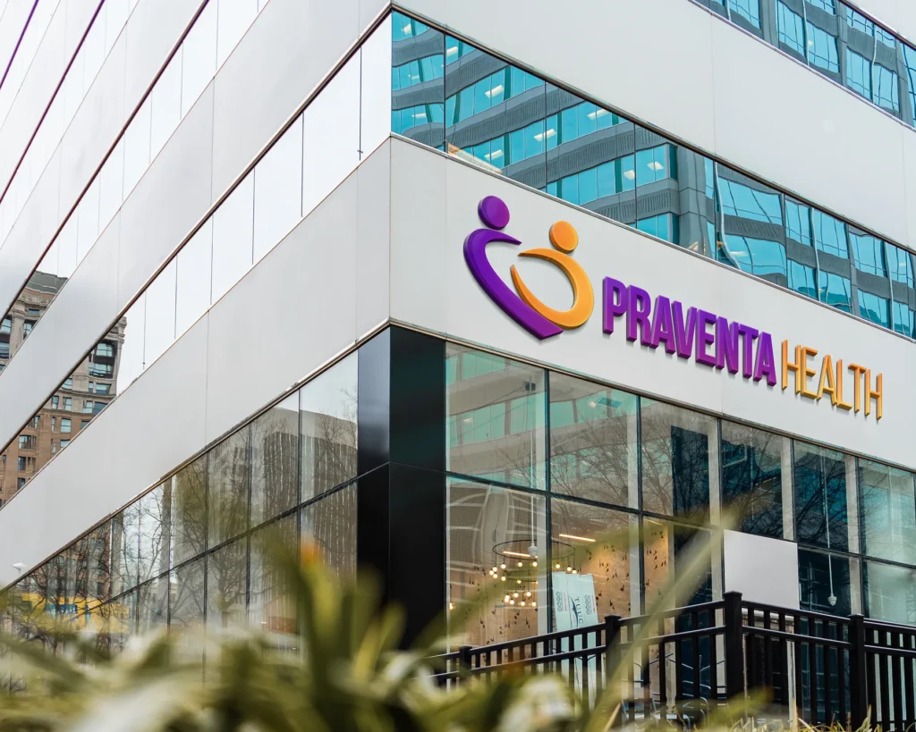 A modern office building with the Praventa Health logo and name displayed on the exterior, featuring purple and gold text and an abstract icon. Reflections of nearby buildings are visible in the glass windows.