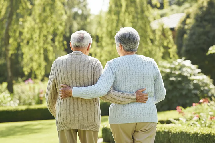 An elderly couple, seen from behind, walks arm in arm through a lush, sunlit garden with green trees and bushes surrounding them, enjoying the health benefits of nature and peaceful moments together.